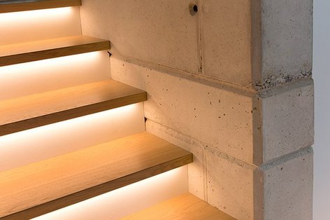 Detailansicht einer beleuchteten Holztreppe mit integrierten LED-Leisten neben einer Sichtbetonwand