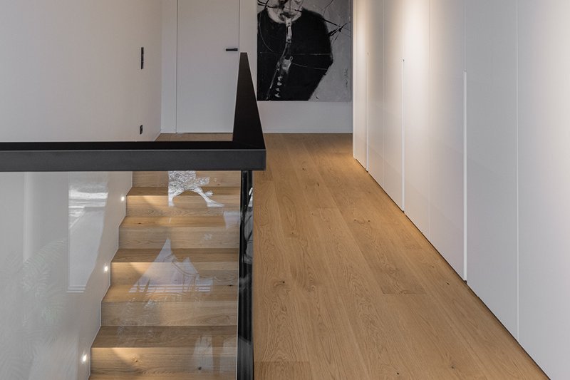 Landhausdiele Eiche astig Natura Flur und Treppenaufgang mit Eichenparkett im Landhausdielen Design. Die Holztreppe ist von einem Glasgeländer mit schwarzem Rand umgeben.