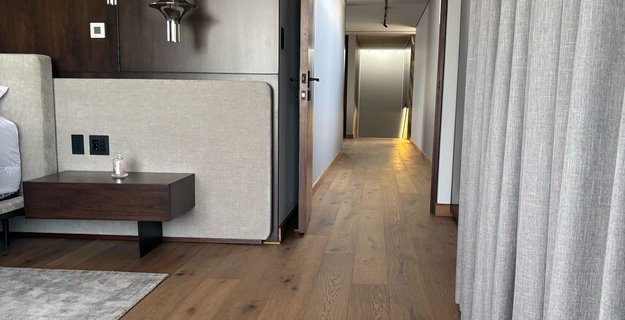 Schlafzimmer mit mittelbraunem Eichenparkett rustikal. Ein beiges Bett, beige Vorhänge und ein grauer Teppich. Eine dunkle Holzwand und chromfarbene Leuchten runden das Raumkonzept ab. Man sieht durch die offene Tür auch in einen Flur, wo auch das Parkett verlegt ist.