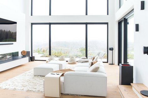 Aussicht in ein modernes Wohnzimmer sowie in die Natur durch große Fenster. der Raum ist in schwarz weiß gehalten und mit einem Holzboden ausgestattet